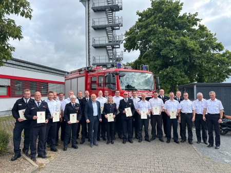 Das Bild zeigt ein Feuerwehrfahrzeug. Im Vordergrund stehen die Kameradinnen und Kameraden mit ihren Ernennungsurkunden. 